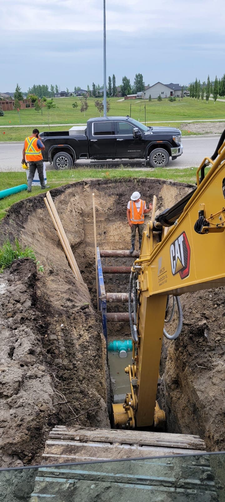 Construction workers and a yellow excavator installing underground pipes in a deep, shored trench.