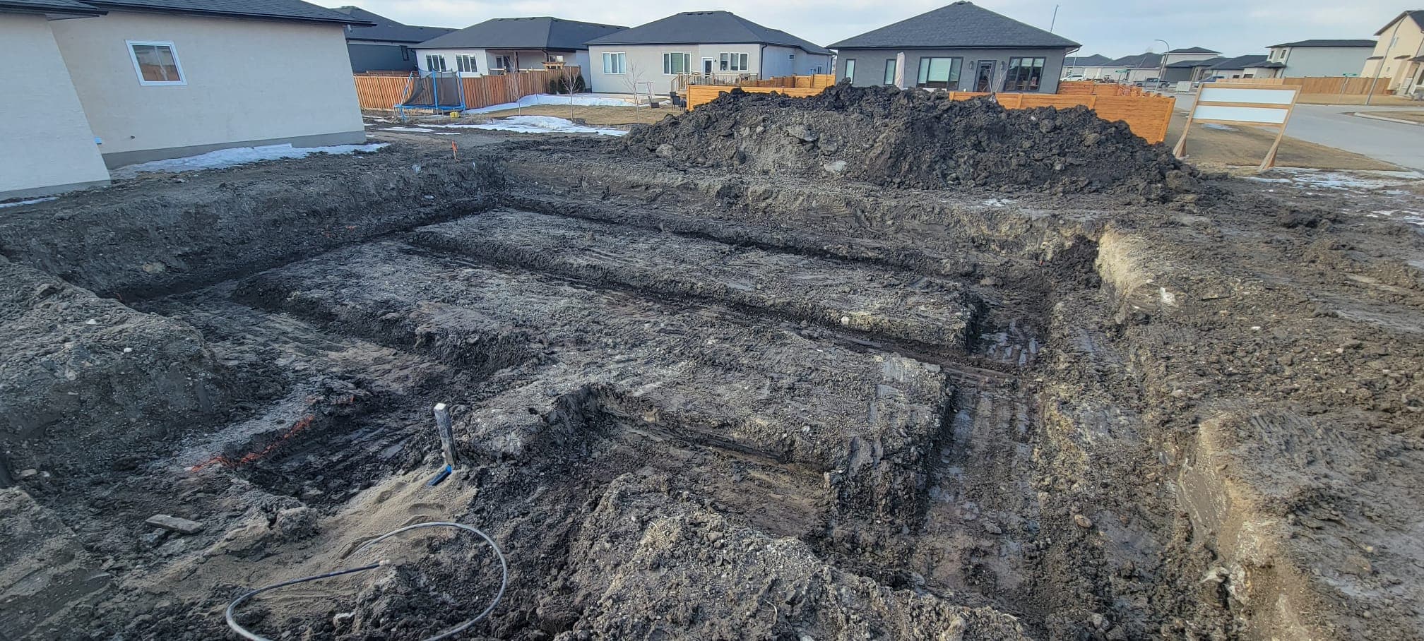 Excavated foundation for a new house in a residential neighborhood with large dirt piles.