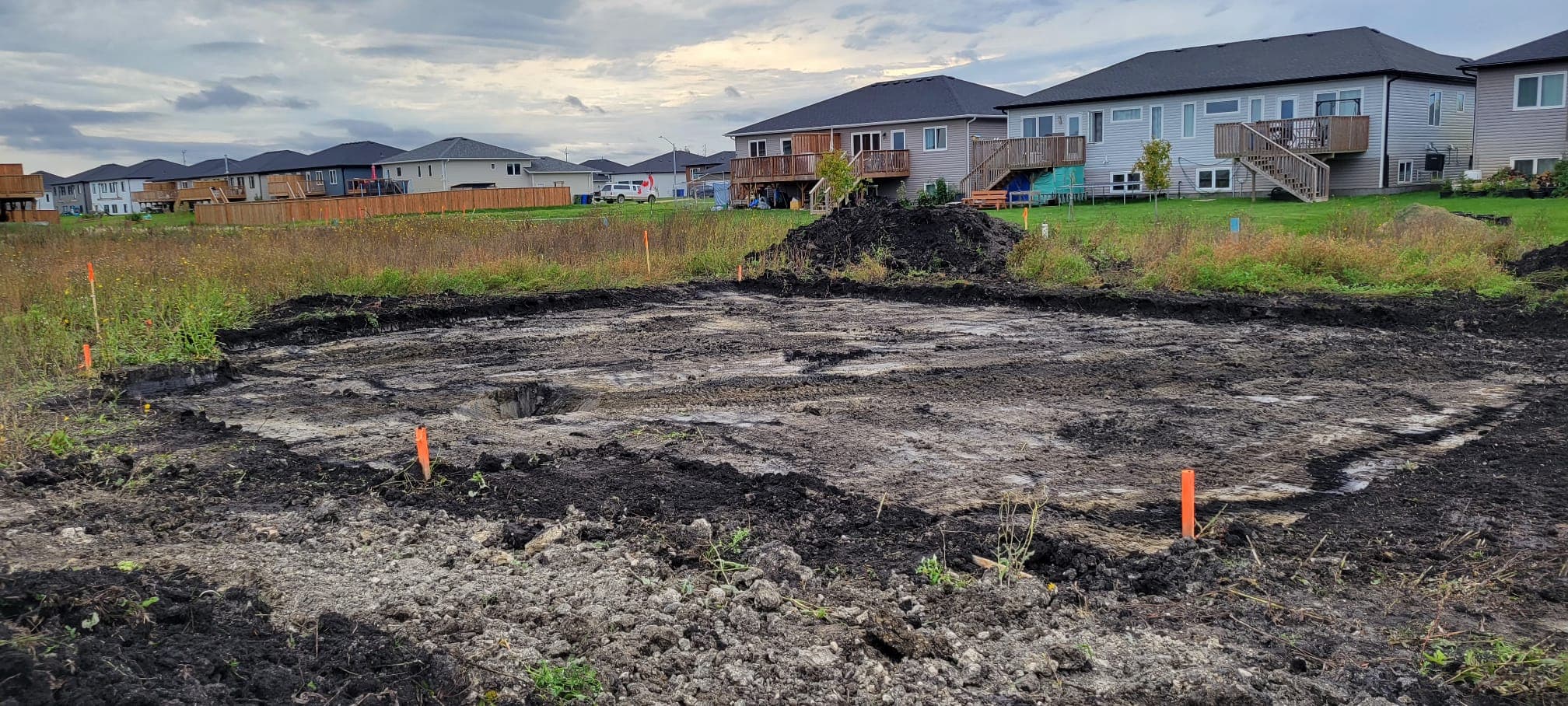 Muddy construction site with orange survey stakes in a suburban neighborhood under a cloudy sky.