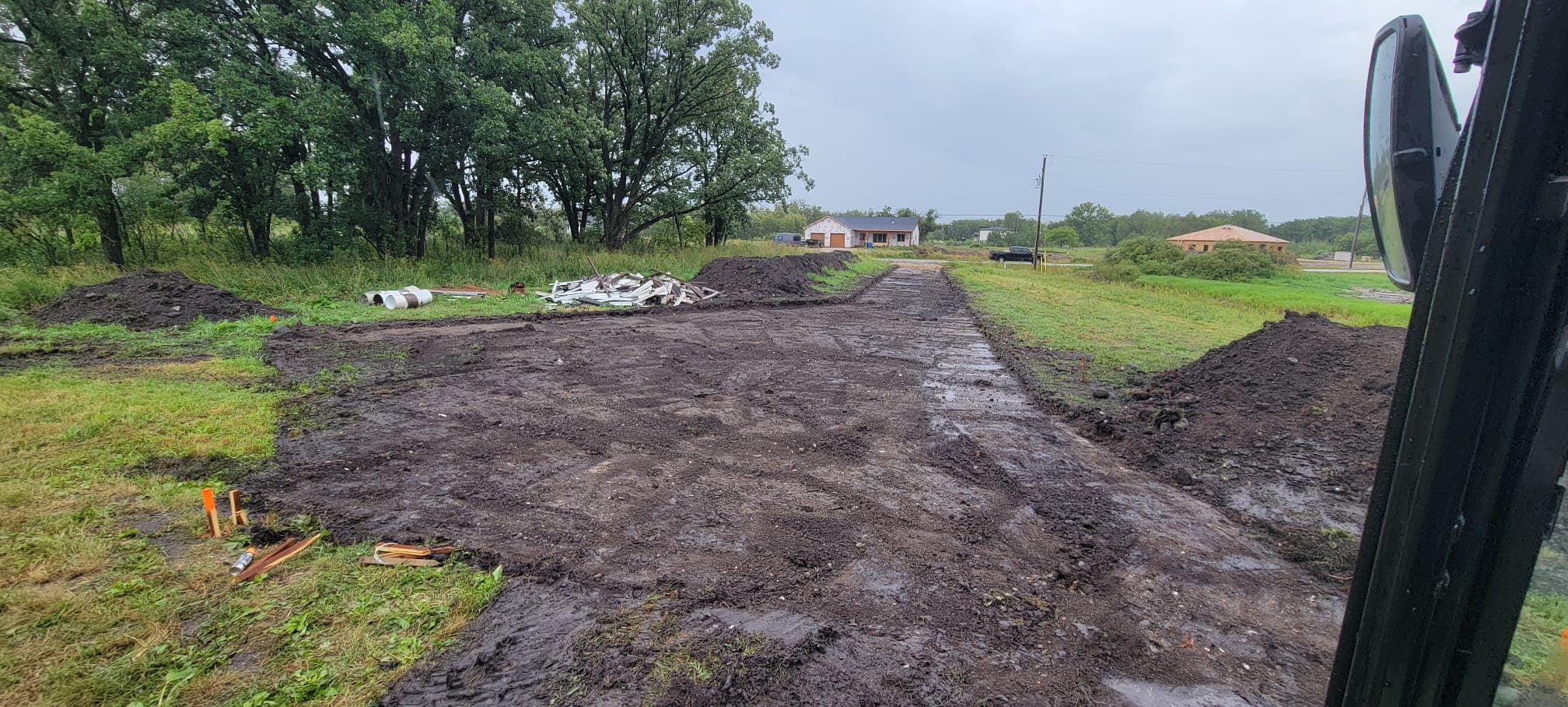 Muddy dirt driveway under construction leading toward houses in a rural, wooded landscape.