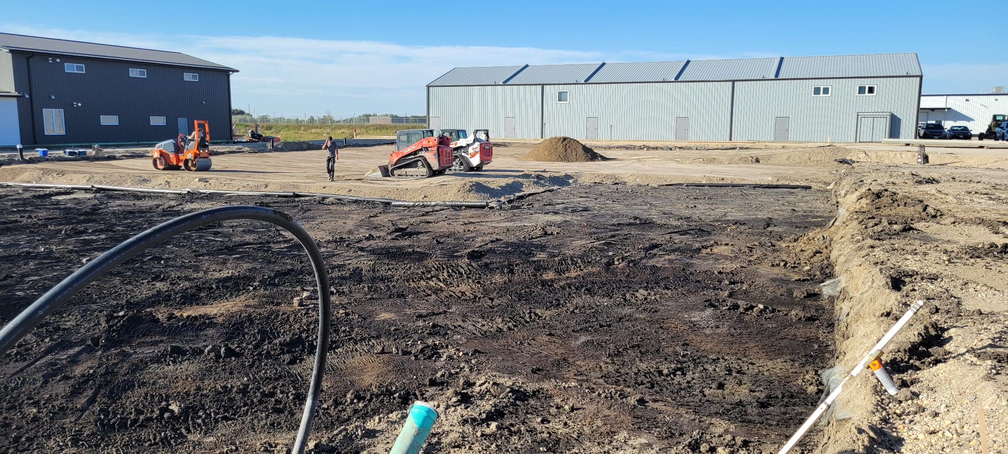 Heavy machinery and workers on a dirt construction site with large industrial buildings behind.