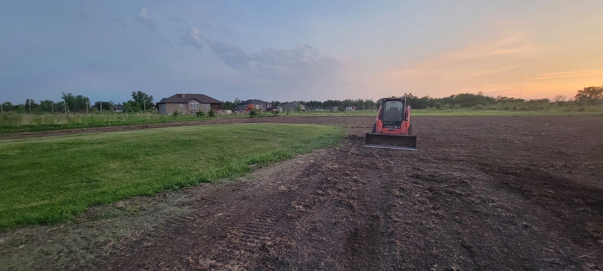 Red skid steer loader on a dirt lot next to a lawn during sunset.