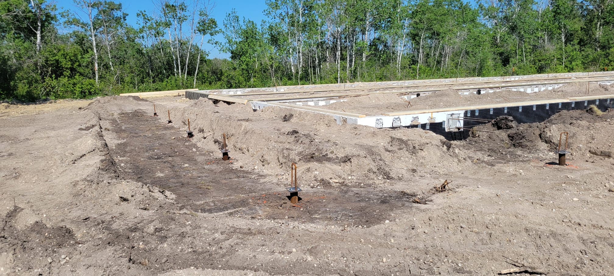 Concrete foundation under construction with metal stakes in the dirt and green trees behind.
